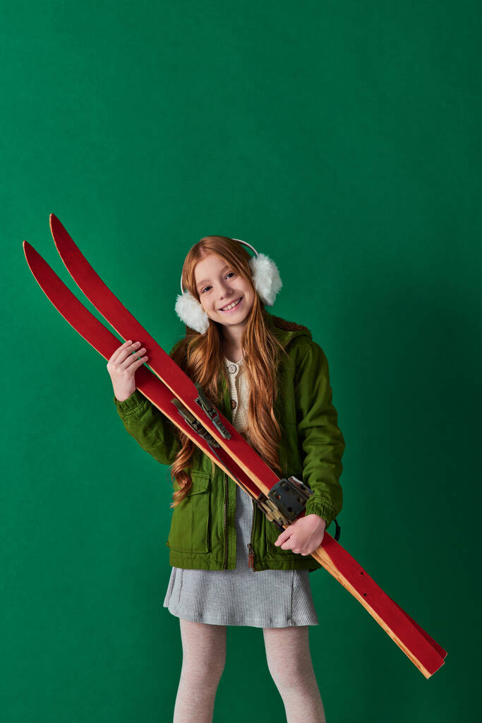 alegre chica preadolescente en orejeras y traje de invierno sosteniendo equipo de esquí rojo sobre fondo turquesa - Foto, imagen