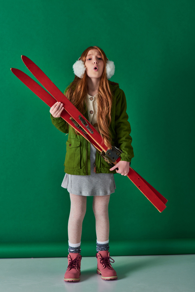 холодный воздух, девочка-подросток в наушниках и зимнем наряде дышит и держит красные лыжи на бирюзовом - Фото, изображение