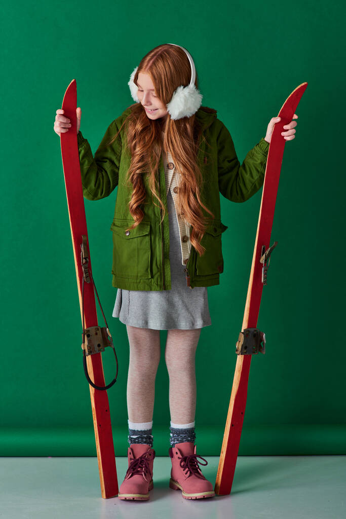 cheerful preteen girl in ear muffs and winter outfit holding red skis on turquoise background - Photo, Image