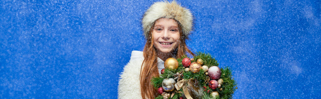 menina feliz em casaco de pele falso e chapéu segurando coroa de Natal decorada sob queda de neve, bandeira - Foto, Imagem