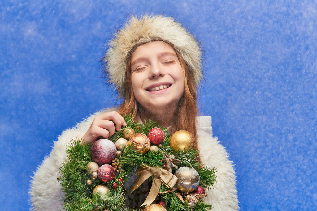 boldog lány csukott szemmel műszőrme kalap és kabát tartja karácsonyi koszorú alá eső hó - Fotó, kép