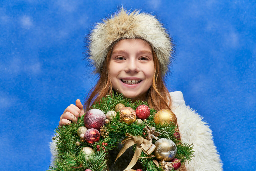 Jahreszeit der Freude, glückliches Mädchen mit Weihnachtskranz unter fallendem Schnee vor blauem Hintergrund - Foto, Bild