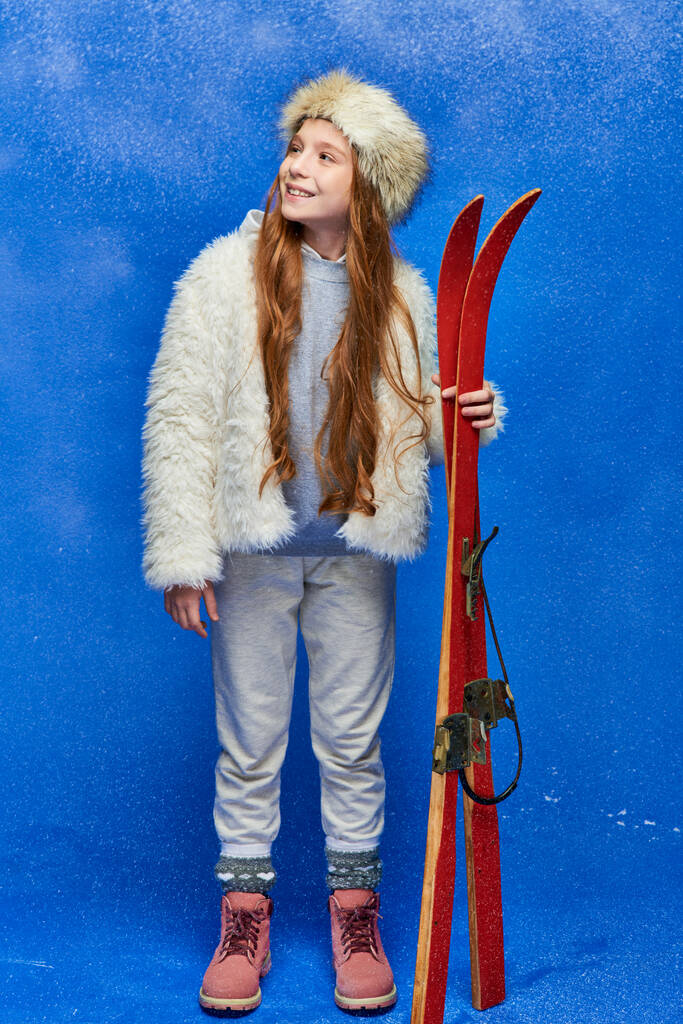 positive preteen girl in winter faux fur jacket and hat holding red skis on turquoise background - Photo, Image