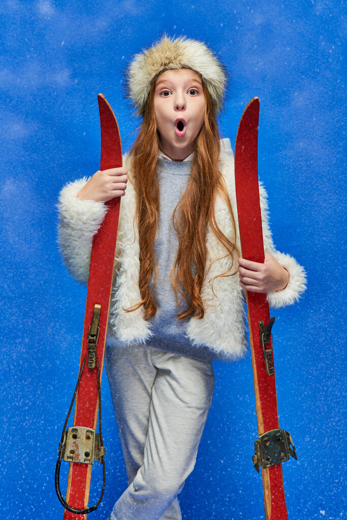 zimní sport, šokované preteen dívka ve falešné kožešinové bundě a klobouk drží lyže na tyrkysovém pozadí - Fotografie, Obrázek