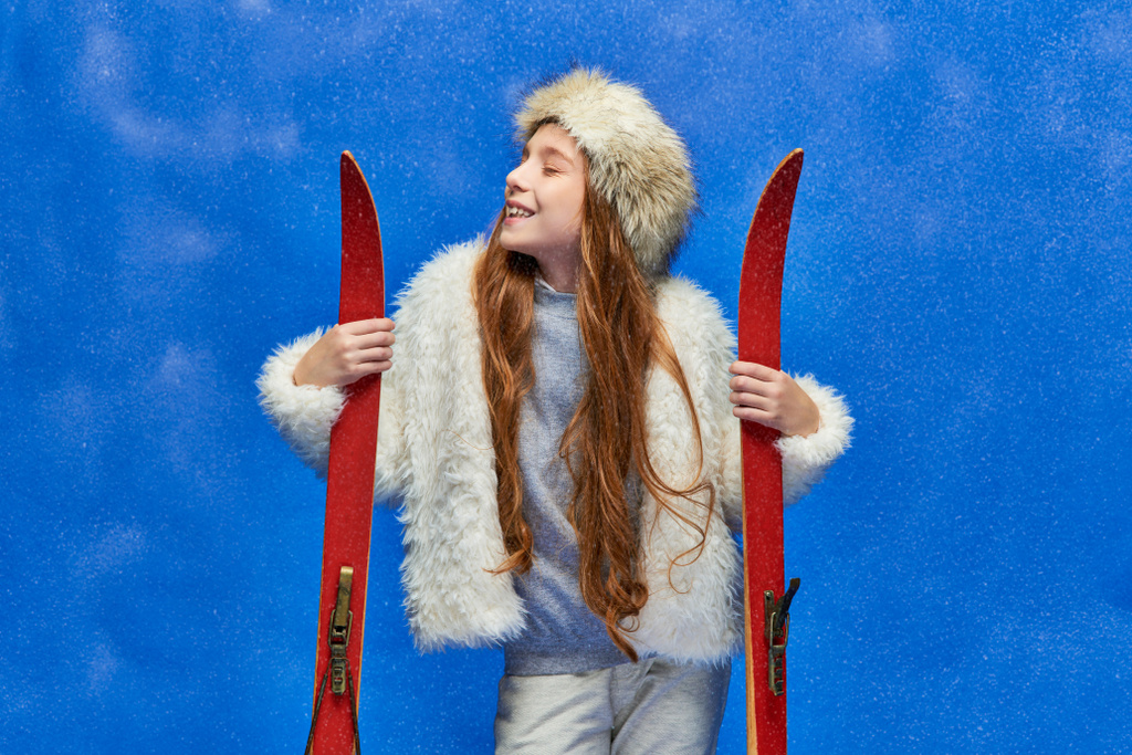 winter joy, pleased preteen girl in faux fur jacket and hat holding red skis on turquoise background - Photo, Image