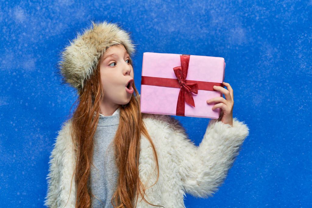 zimní dovolená, šokovaná dívka ve falešné kožešinové bundě a klobouku drží dárkovou krabici na tyrkysovém pozadí - Fotografie, Obrázek