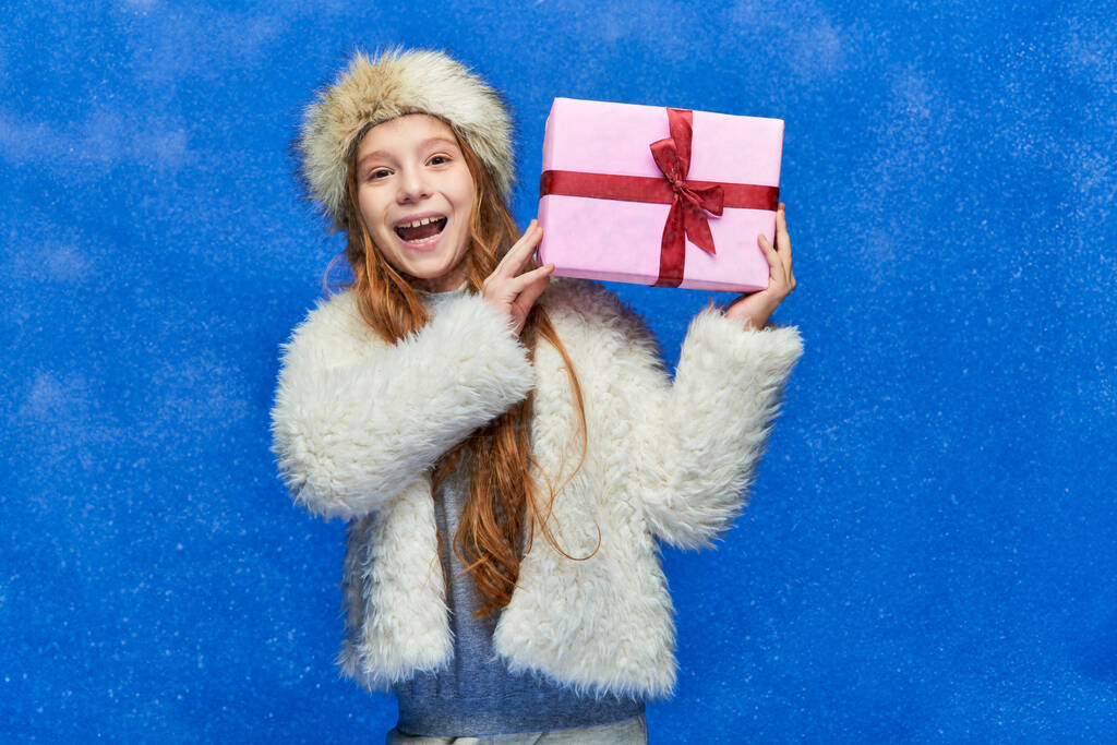 winter holidays, cheerful girl in faux fur jacket and hat holding gift box on turquoise background - Photo, Image