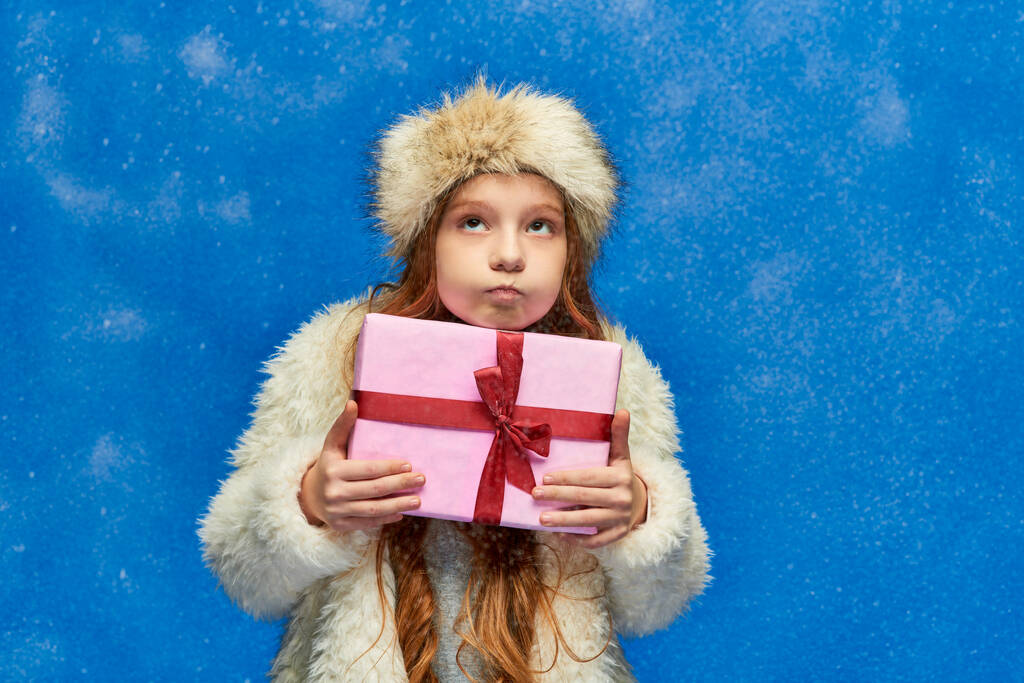 férias de inverno, menina em casaco de pele falso e bochechas inchadas chapéu e segurando caixa de presente em turquesa - Foto, Imagem