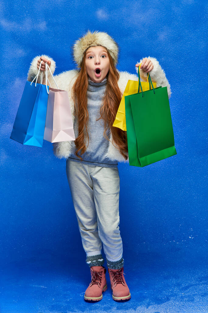 holiday shopping, shocked girl in faux fur jacket and hat holding shopping bags on turquoise, snow - Photo, Image