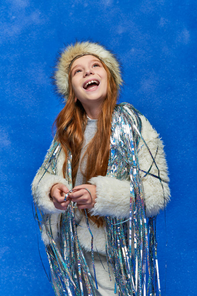 fille excitée en fausse veste de fourrure et chapeau avec tinsel debout sous la neige tombante sur fond bleu - Photo, image