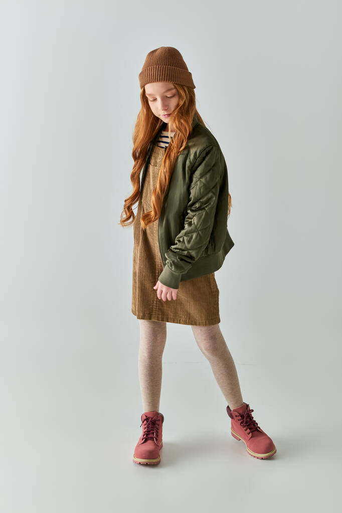 милая девочка-подросток с длинными волосами, стоящими в шляпе, платье и зимняя куртка и сапоги на сером фоне - Фото, изображение