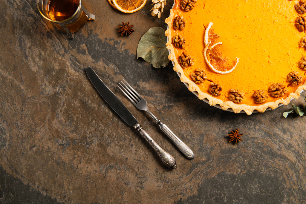 тыквенный пирог с грецкими орехами и апельсиновыми ломтиками рядом со старинными столовыми приборами и теплым чаем, в обстановке Дня благодарения - Фото, изображение