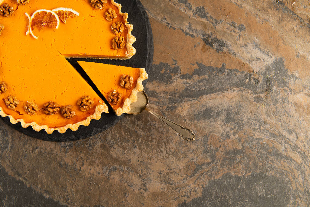 torta espátula cerca de pastel de acción de gracias adornado con rodajas de naranja y nueces en mesa de piedra texturizada - Foto, imagen