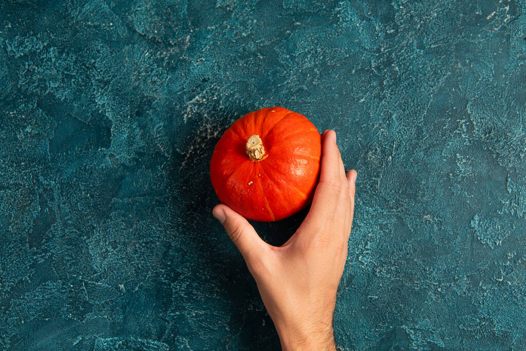 vue recadrée de la main masculine près de citrouille orange vif sur la surface texturée bleue, concept d'action de grâce - Photo, image