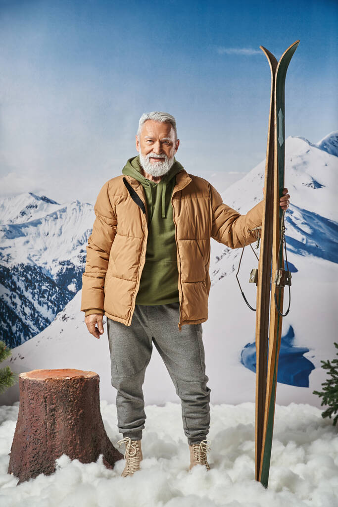 Santa deportivo alegre con barba blanca sosteniendo esquí con montañas nevadas en el telón de fondo, concepto de invierno - Foto, imagen