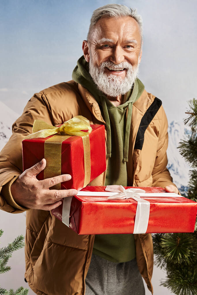 hombre alegre vestido como Santa sosteniendo regalos rojos y sonriendo sinceramente a la cámara, concepto de invierno - Foto, Imagen