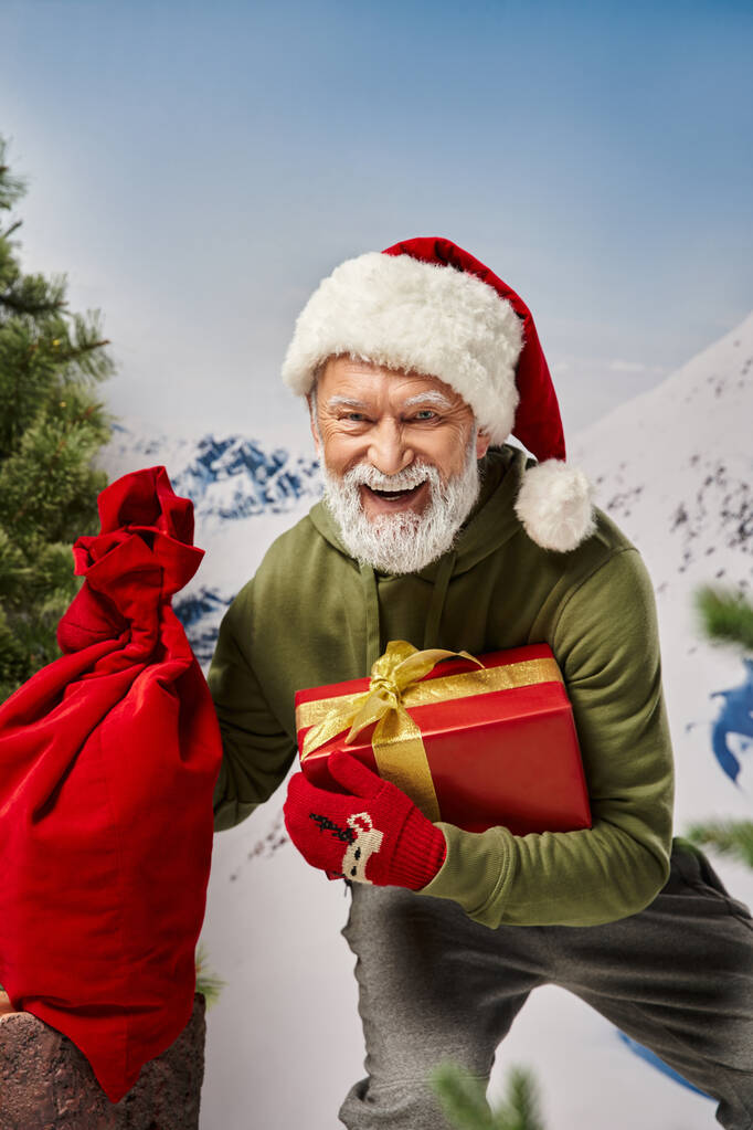 jolly Santa with white beard in christmassy hat with present bag and gift in hand, winter concept - Photo, Image