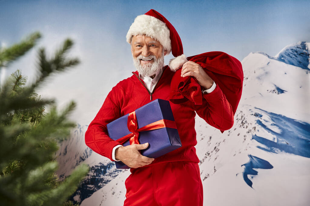 joyful Santa in red attire smiling at camera with present and gift bag in hands, winter concept - Photo, Image