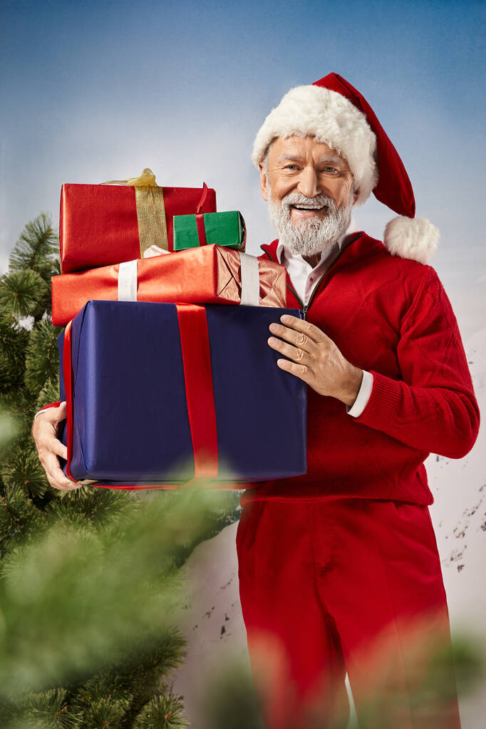 Kırmızı Noel Baba kostümlü neşeli adam elinde hediyelerle kameraya gülümsüyor, kış konsepti. - Fotoğraf, Görsel