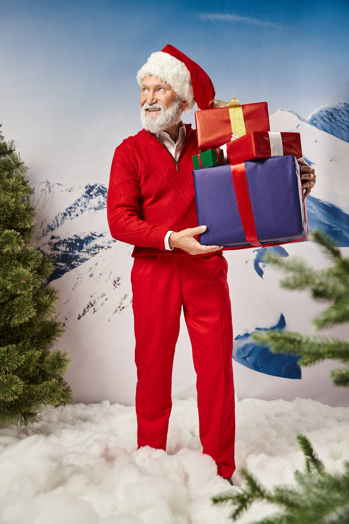 smiley bearded Santa with presents in hands looking away with mountain backdrop, winter concept - Photo, Image