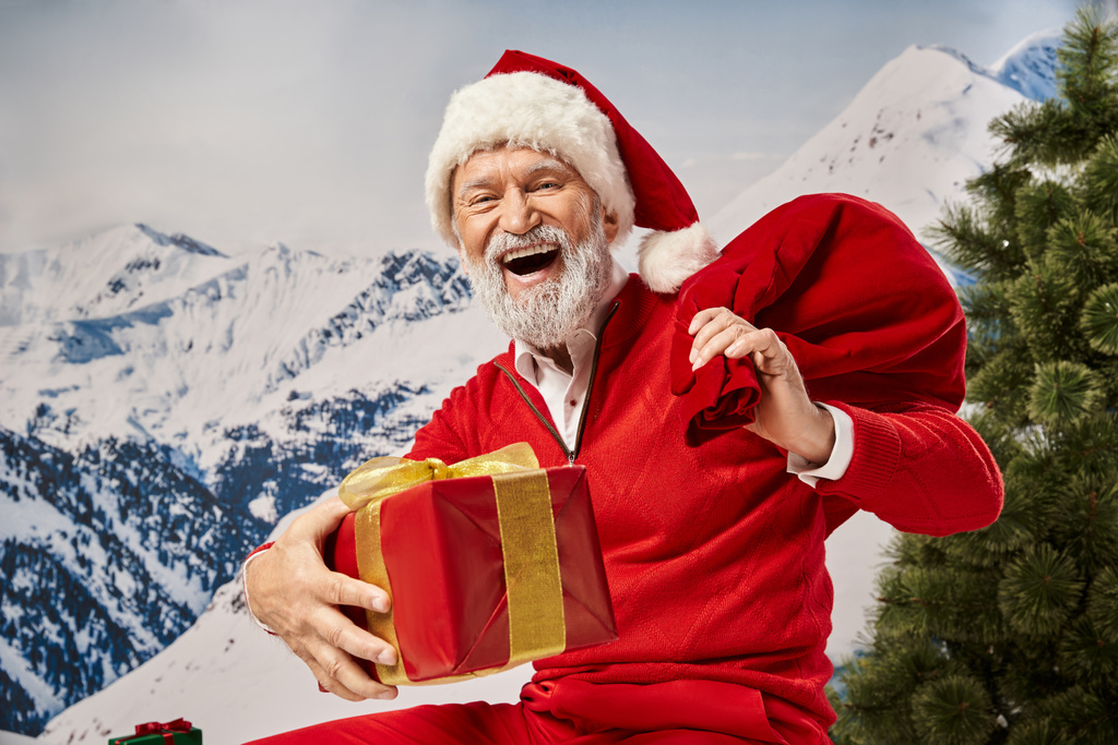весёлый человек, одетый как Санта, счастливо улыбается перед камерой, держа подарочную сумку и подарок, зимняя концепция - Фото, изображение