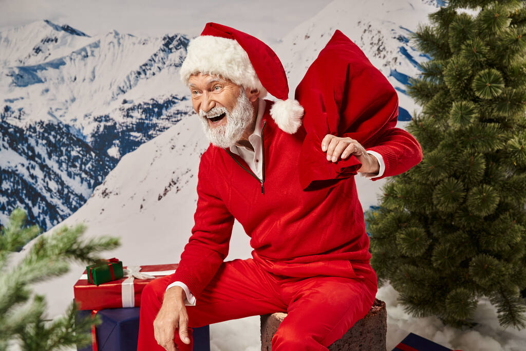 hombre alegre en traje de Santa riendo y sentado en el tocón del árbol con bolsa de regalo, Feliz Navidad - Foto, Imagen