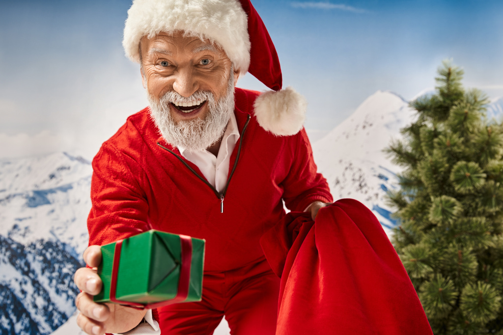 radosny człowiek przebrany za Mikołaja pożycza prezent przed kamerą i uśmiecha się wesoło, koncepcja zimowa - Zdjęcie, obraz