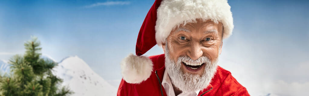 cheerful Santa in hat smiling happily at camera with mountain backdrop, winter concept, banner - Photo, Image