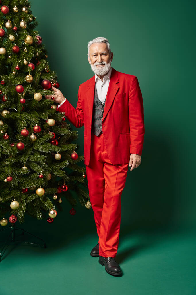 stylový pohledný Santa Claus pózující poblíž vánoční stromeček dotýkající se dekorace, zimní koncept - Fotografie, Obrázek