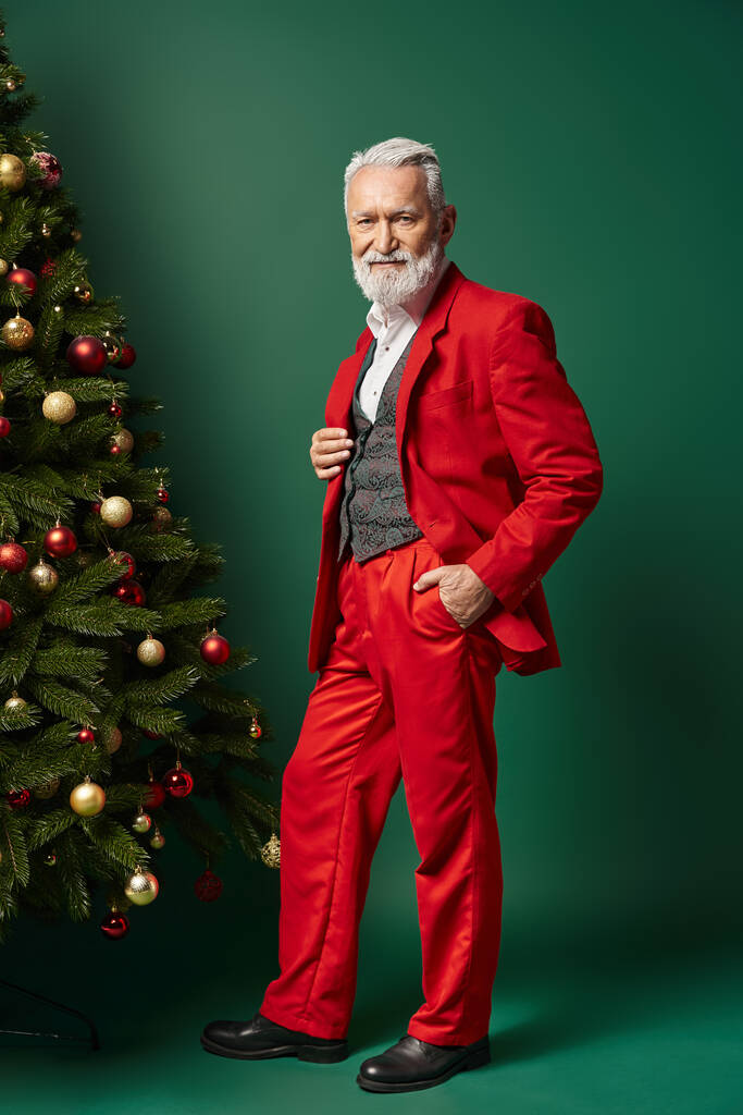 elegantní Santa v červeném obleku s bílým plnovousem pózuje poblíž jedle s rukou v kapse, zimní koncept - Fotografie, Obrázek