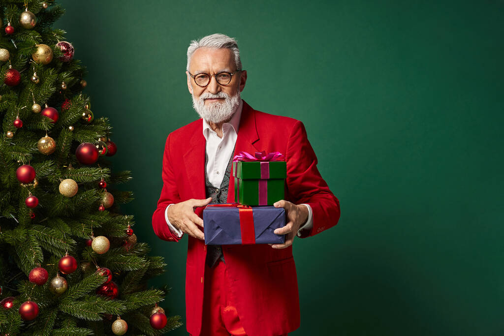élégant Père Noël avec lunettes et barbe tenant pile de cadeaux sur fond vert foncé, concept d'hiver - Photo, image