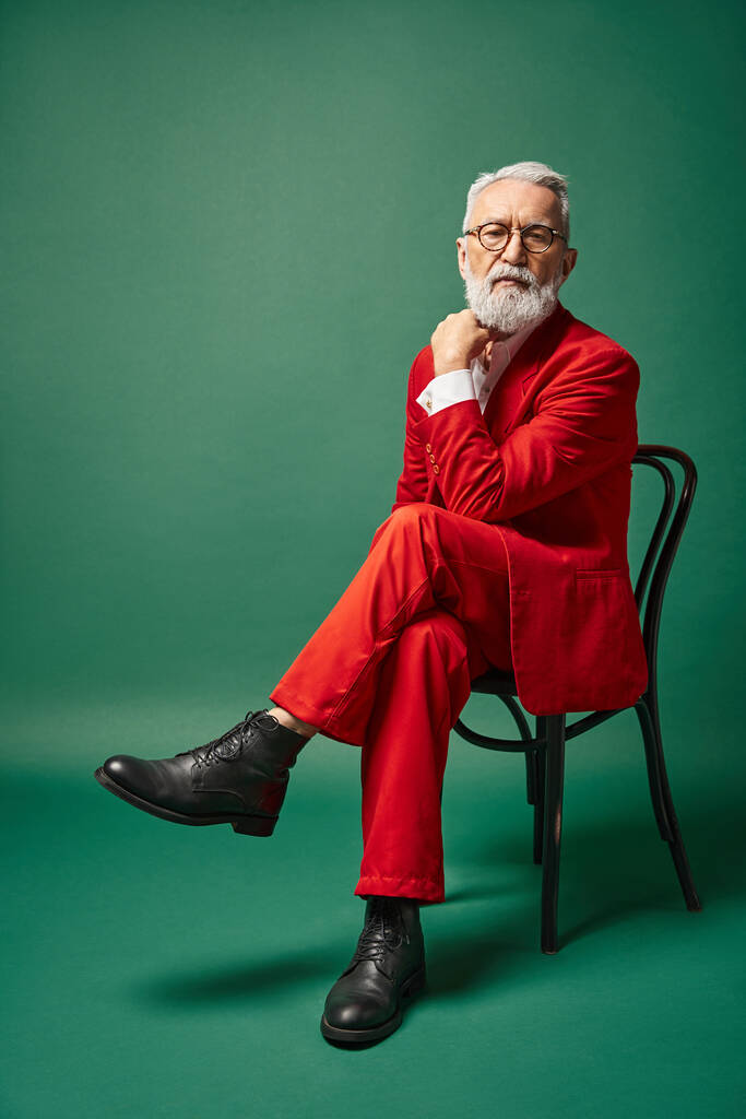 stijlvolle man verkleed als kerstman met bril zittend op stoel met de hand in de buurt gezicht, winter concept - Foto, afbeelding