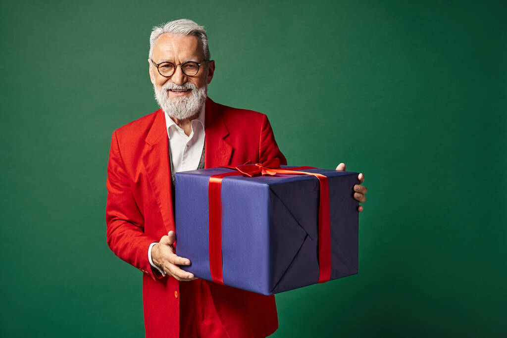 elegant Santa with beard and glasses holding huge present and smiling sincerely, winter concept - Photo, Image