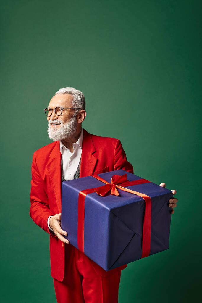 stylový elegantní Santa drží obrovský dárek na zeleném pozadí a odvrací pohled, zimní koncept - Fotografie, Obrázek