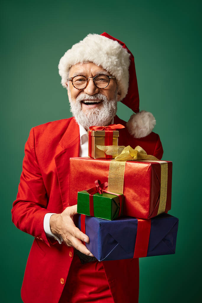 portrait of elegant Santa in red attire holding bunch of presents smiling at camera, winter concept - Photo, Image