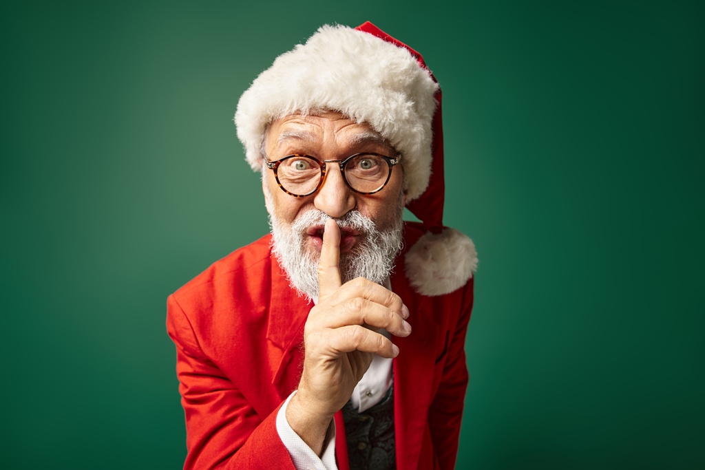 stylish Santa in red hat and glasses showing silence gesture looking at camera, winter concept - Photo, Image