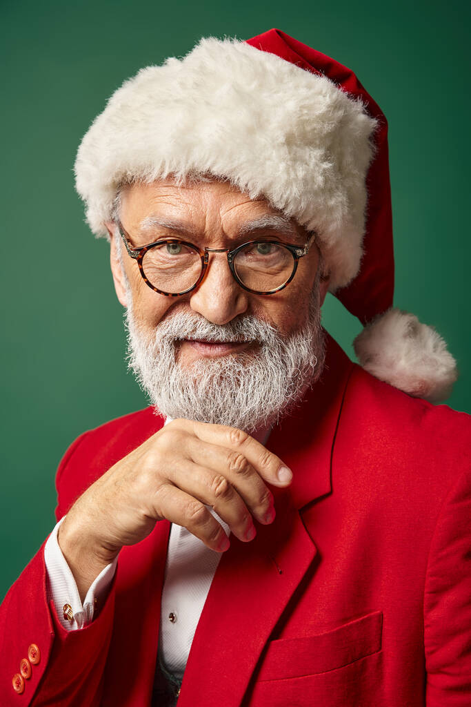 elegante Santa con sombrero rojo y gafas mirando a la cámara con la mano bajo la barbilla, concepto de invierno - Foto, Imagen