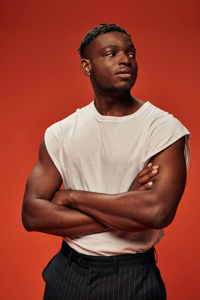 νεαρός αφροαμερικάνος με αυτοπεποίθηση, με λευκό μπλουζάκι, κοιτάζει αλλού με διπλωμένα χέρια στο κόκκινο. - Φωτογραφία, εικόνα