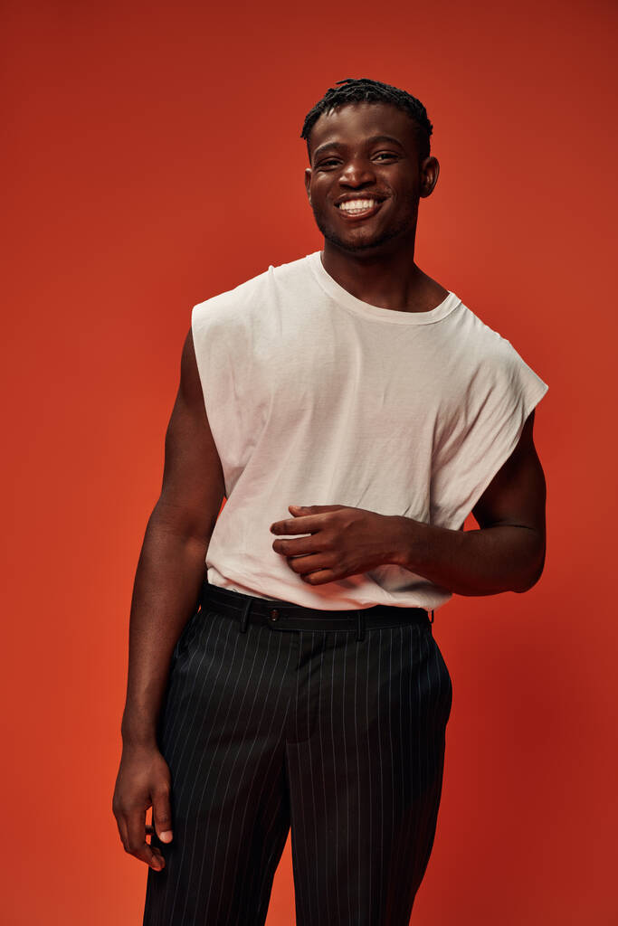 veselý africký Američan v bílém tank top a černé kalhoty při pohledu na kameru na červeném pozadí - Fotografie, Obrázek