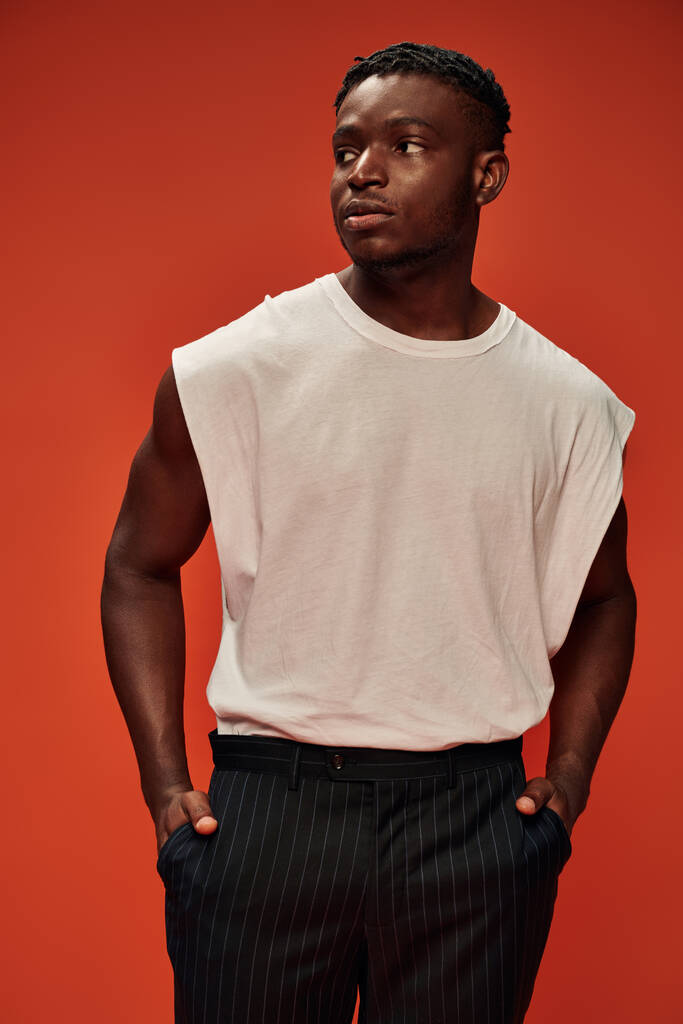 sebevědomý a stylový africký americký mužský model, který se drží za ruce v kapsách a dívá se jinam na červenou - Fotografie, Obrázek