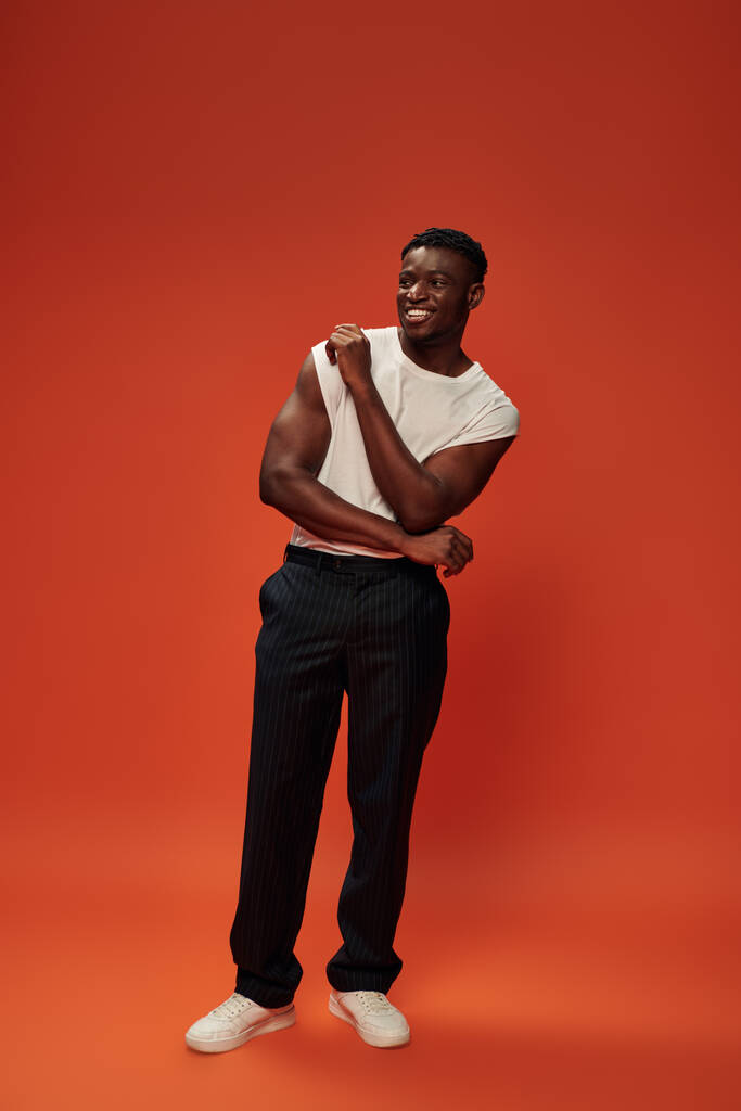 piena lunghezza di gioioso afro americano uomo in pantaloni neri in piedi su sfondo rosso e arancione - Foto, immagini