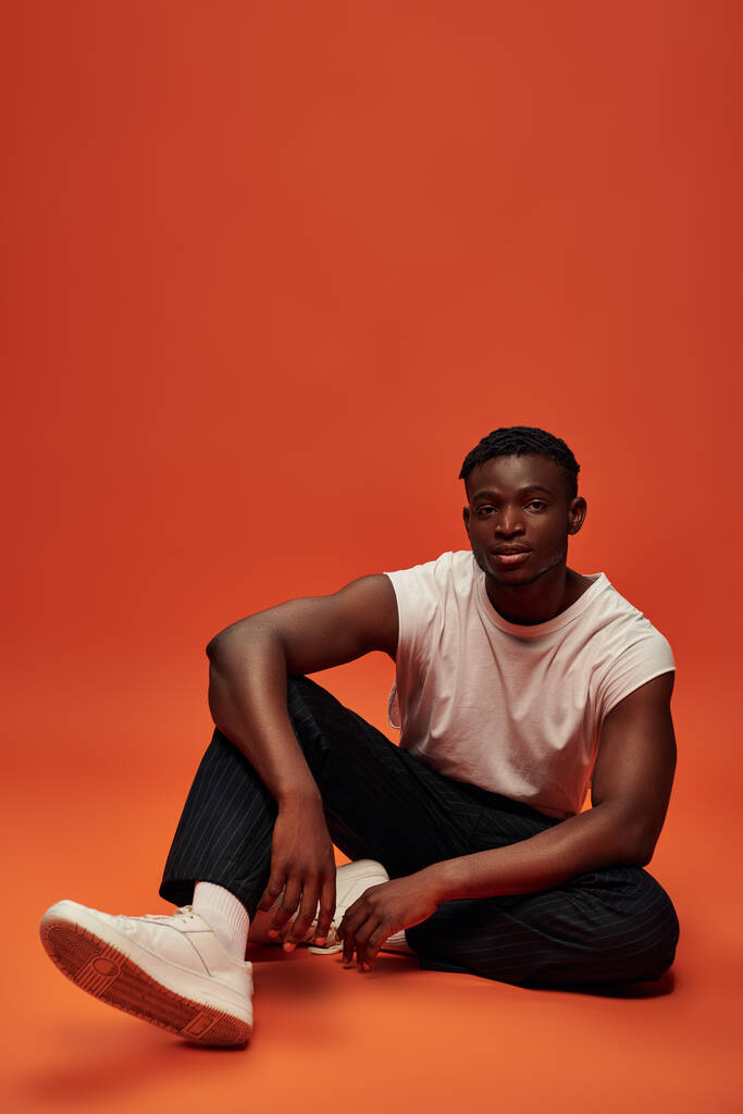 афроамериканський чоловік у модному повсякденному одязі сидить і дивиться на камеру на червоному і помаранчевому тлі - Фото, зображення