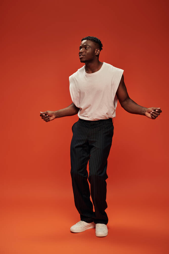 asombrado y sonriente hombre afroamericano en ropa de moda mirando hacia otro lado en el fondo rojo y naranja - Foto, imagen