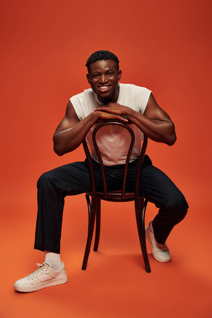 podekscytowany Afroamerykanin człowiek w białym zbiorniku top i spodnie siedzi na krześle na czerwonym i pomarańczowym tle - Zdjęcie, obraz