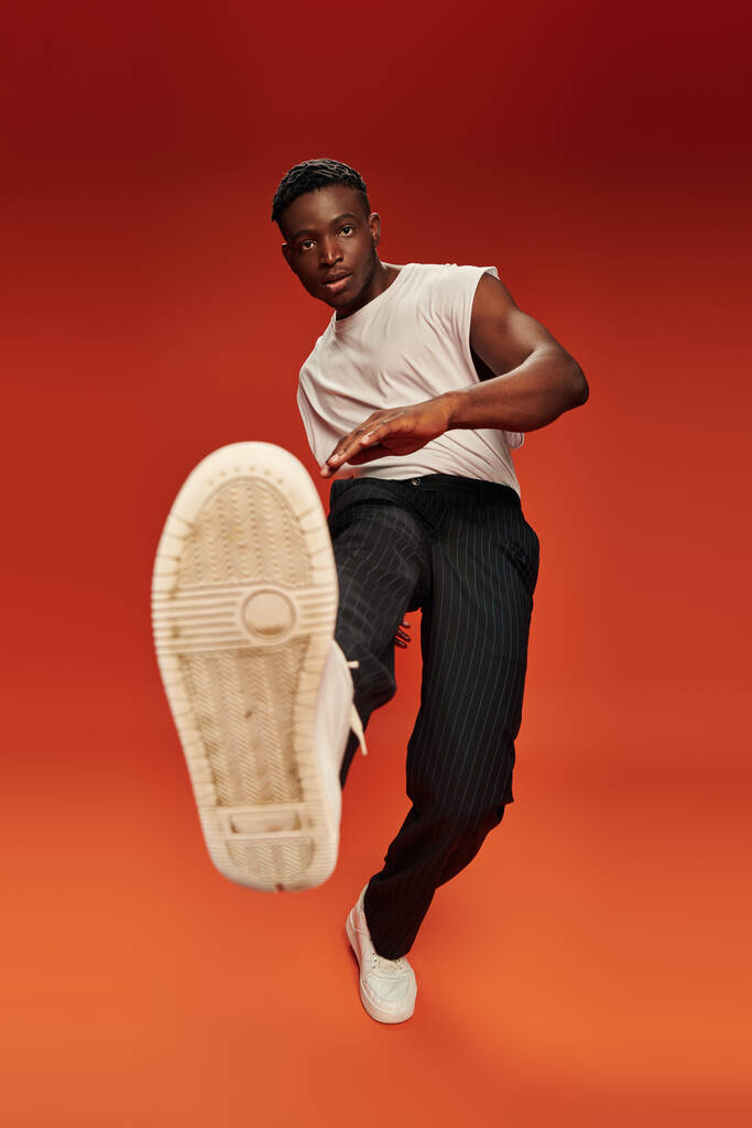 stylový africký Američan v odvážné póze s nataženou nohou na červeném a oranžovém pozadí - Fotografie, Obrázek