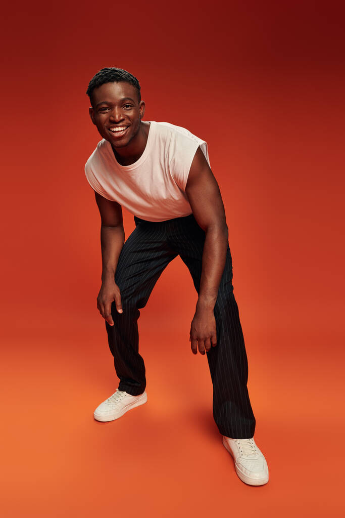 longitud completa de hombre afroamericano de moda y feliz riéndose de la cámara en el fondo rojo y naranja - Foto, imagen