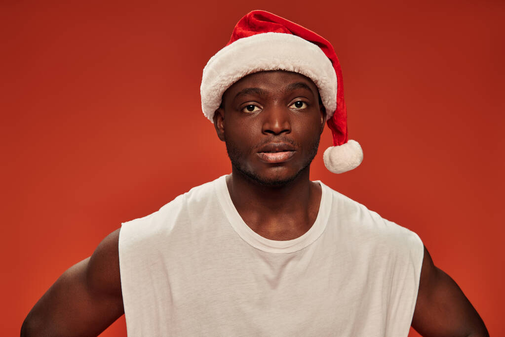 vakava afrikkalainen amerikkalainen mies valkoinen toppi ja jouluhattu katsot kameraa punaisella taustalla - Valokuva, kuva