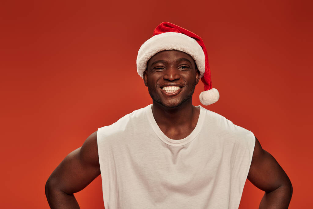vrolijk Afrikaans amerikaanse man met stralende glimlach poseren in santa hoed en kijken naar de camera op rood - Foto, afbeelding