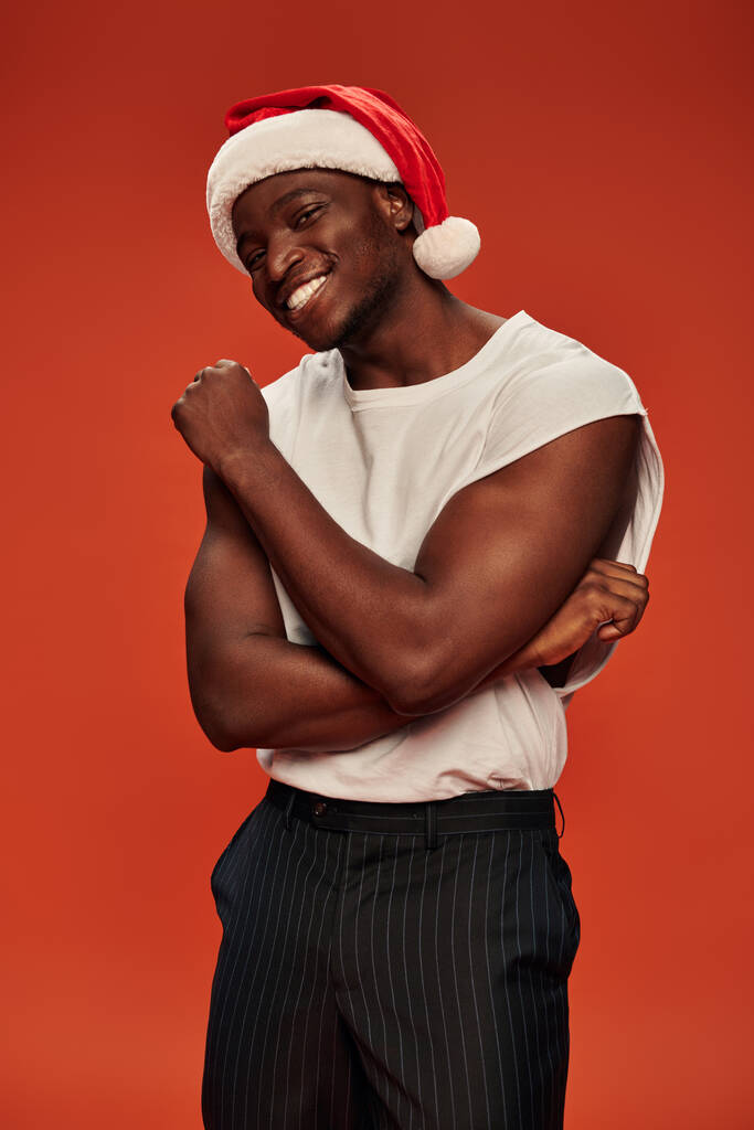 caldo e atletico ragazzo africano americano in cappello di Babbo Natale e canotta bianca sorridente alla fotocamera sul rosso - Foto, immagini