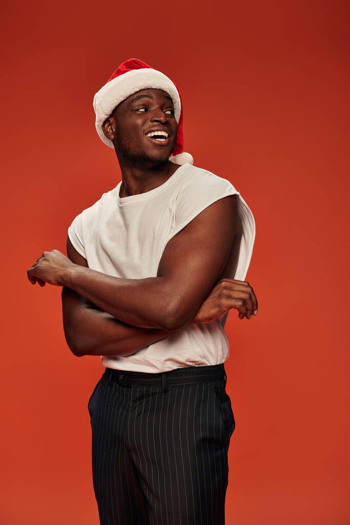 hombre afroamericano emocionado en camiseta blanca y gorra de Navidad mirando hacia otro lado en el fondo rojo - Foto, imagen